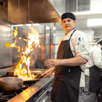 Smiling male student wearing kitchen cap and apron looks at the camera while holding a pan over the stove, a large flame jumping up out of the pan..